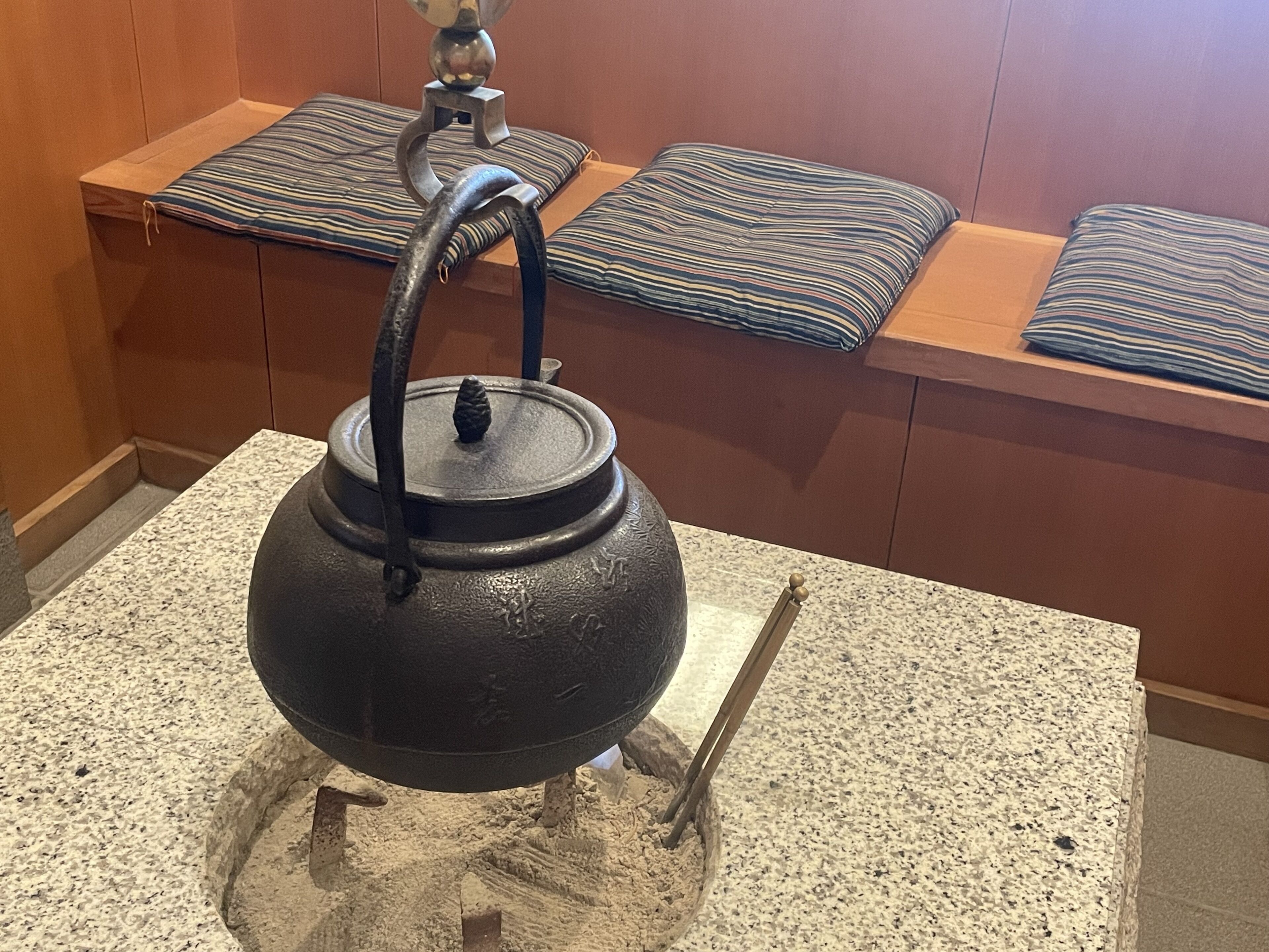 Una tetera negra tradicional sobre una encimera de granito, con un banco de madera y cojines a rayas al fondo.