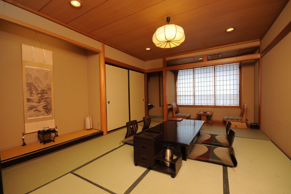 Ein traditioneller japanischer Raum mit Tatami-Boden, einem niedrigen Tisch, Stühlen, einer Schiebetür und einem großen Fenster mit Ausblick.