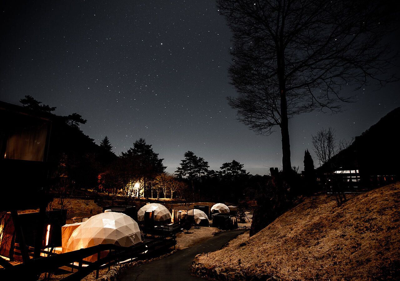 A starry night sky with several dome-shaped tents illuminated by warm lights, set against a backdrop of trees and a hill.