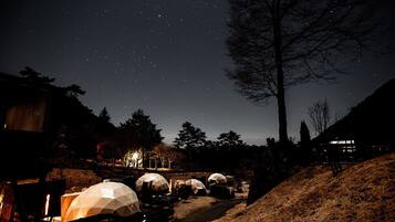 A starry night sky with several dome-shaped tents illuminated by warm lights, set against a backdrop of trees and a hill.