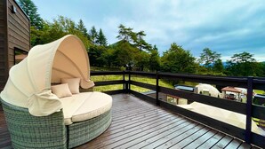 Una silla redonda de mimbre con dosel en una terraza de madera, con vistas a una zona ajardinada con árboles y un edificio al fondo.