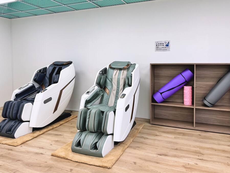 Two massage chairs in a room with a wooden floor, a shelf with yoga mats, and a sign on the wall.