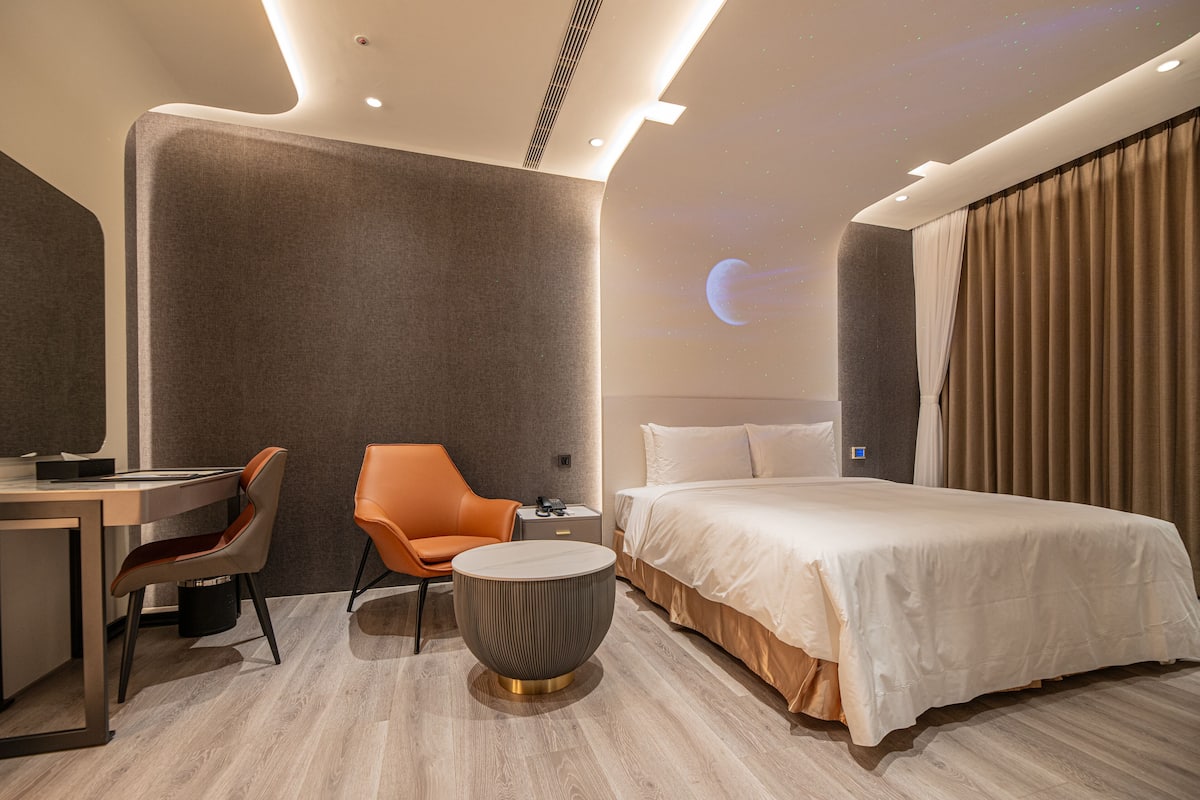 A modern hotel room with a bed, a desk, an orange chair, and a round coffee table.