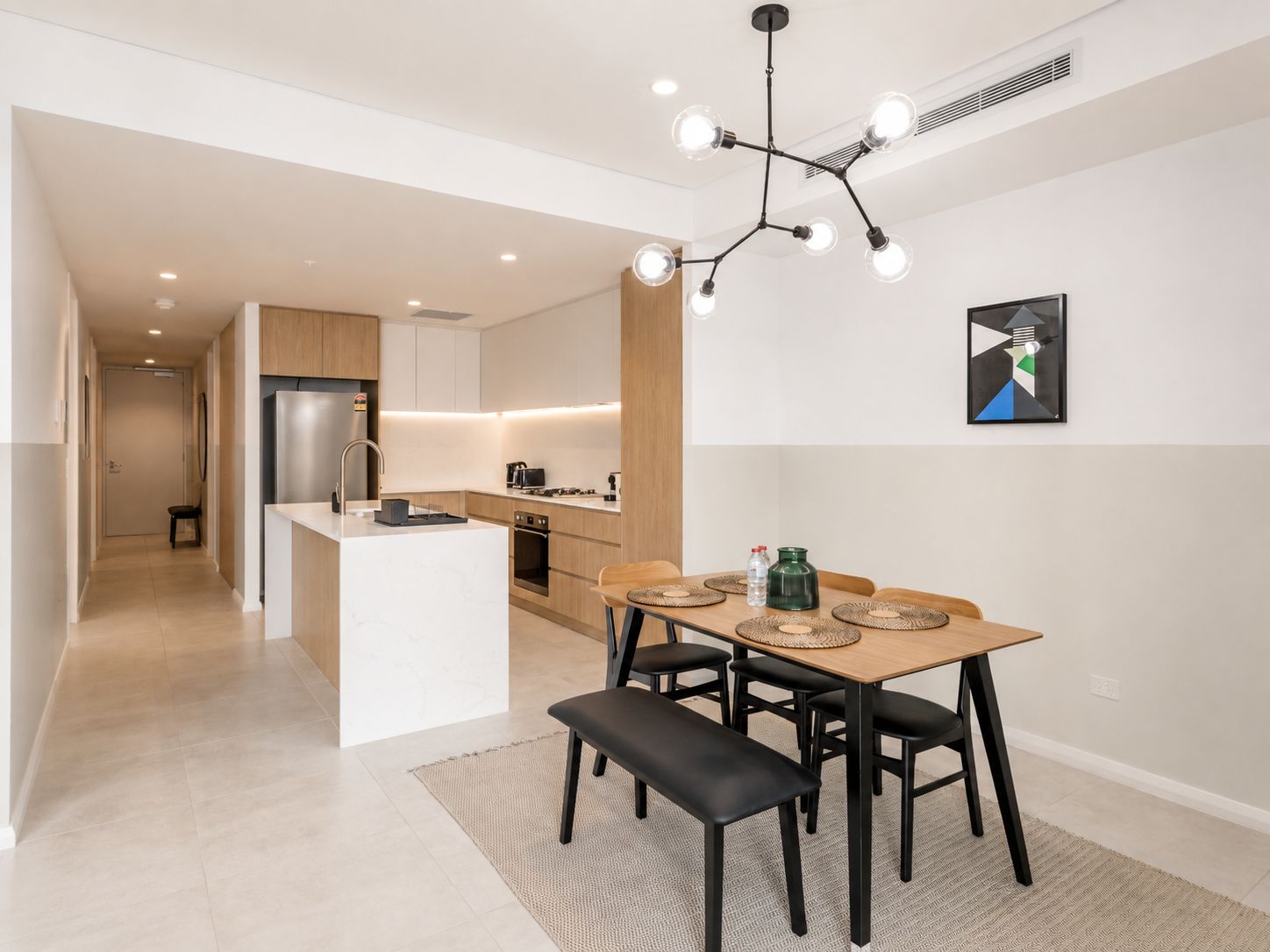 Una cocina moderna con una isla central, un comedor con mesa y sillas de madera, y una lámpara contemporánea.