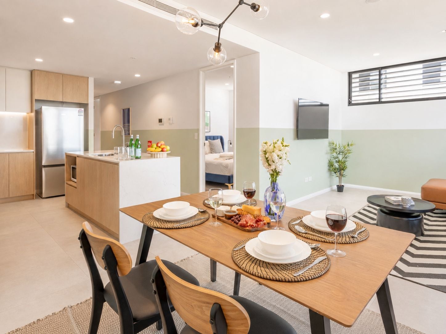 A modern kitchen with a dining area, a living space with a sofa and a TV, and a small balcony with a potted plant.