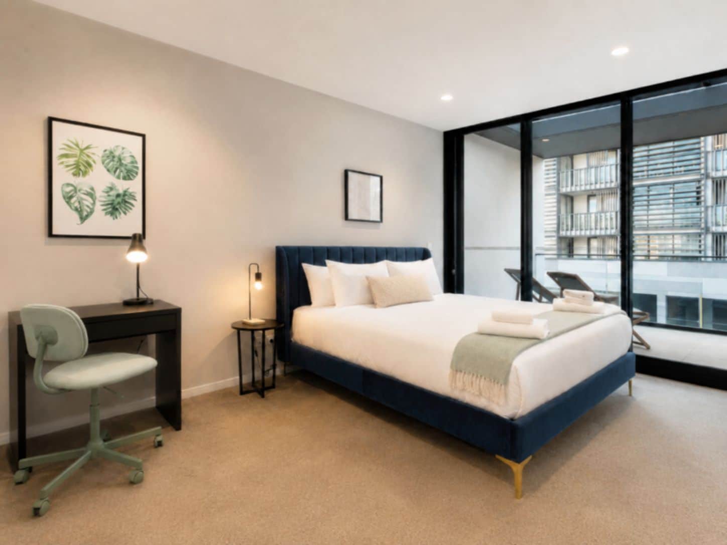 A modern hotel room with a large bed, a desk, a chair, and a view of the city through floor-to-ceiling windows.