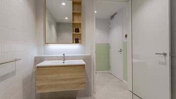 Un baño moderno con pared de azulejos blancos, tocador de madera con lavabo, un espejo y una puerta que da a otra habitación.