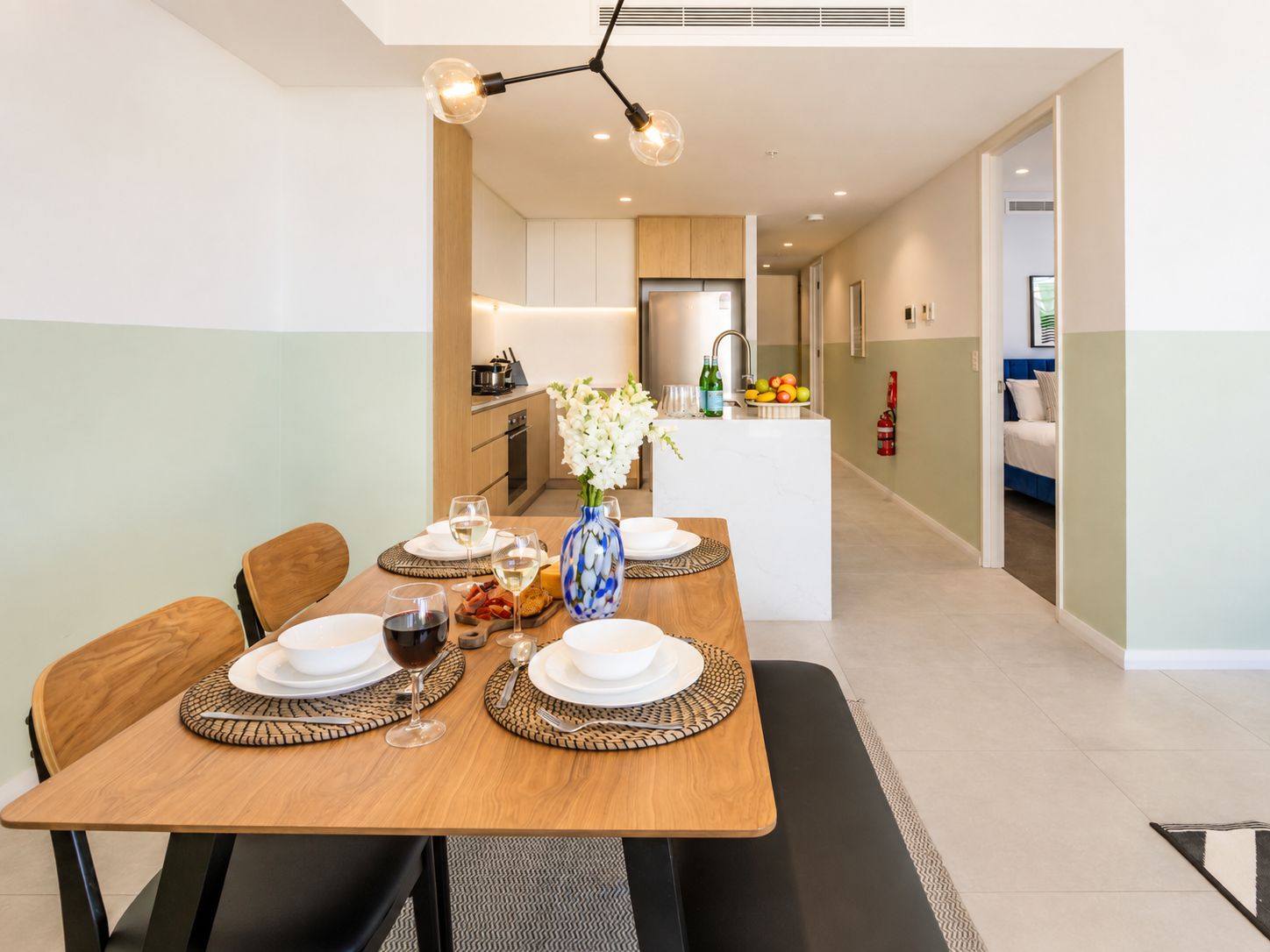 A modern dining area with a wooden table set for four, a vase with flowers, and a kitchen in the background.