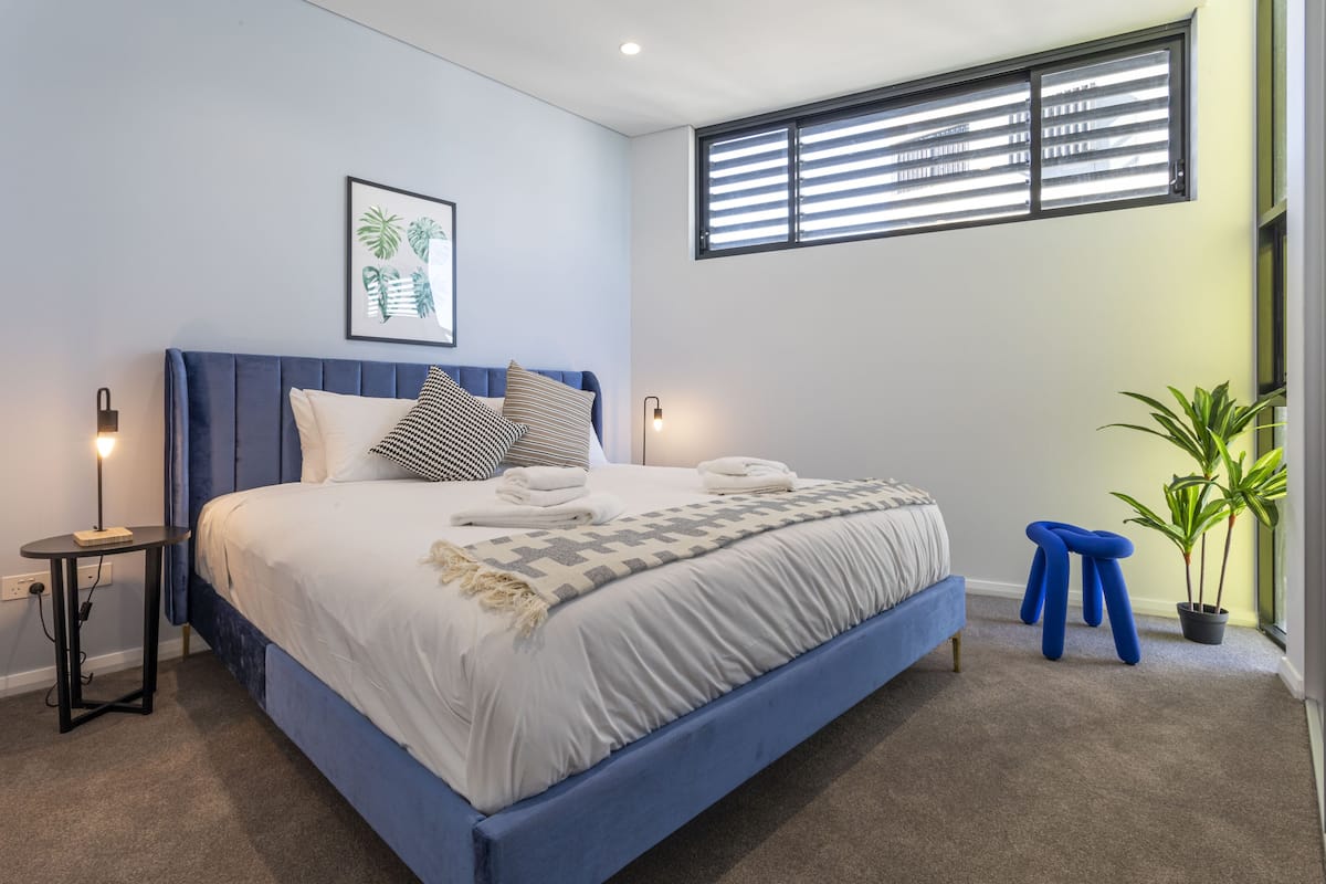 Un dormitorio con una cama grande, una mesita de noche, un cuadro en la pared, una ventana con persianas y una planta en maceta.