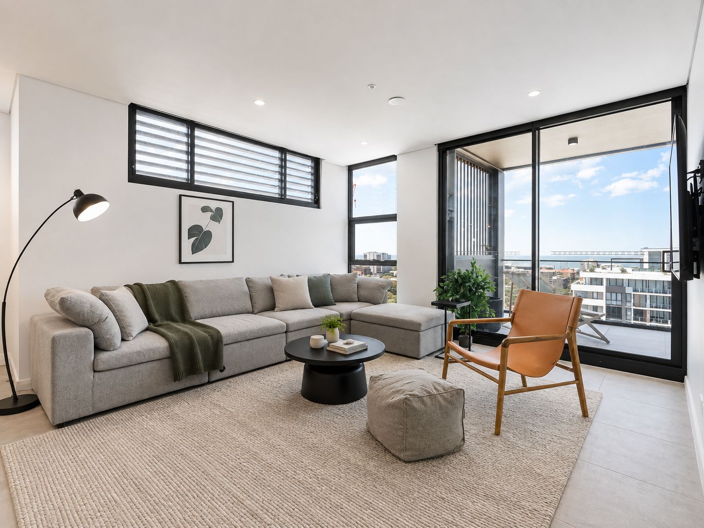 A modern living room with a sectional sofa, a round coffee table, a floor lamp, a single chair, and a large window offering a city view.