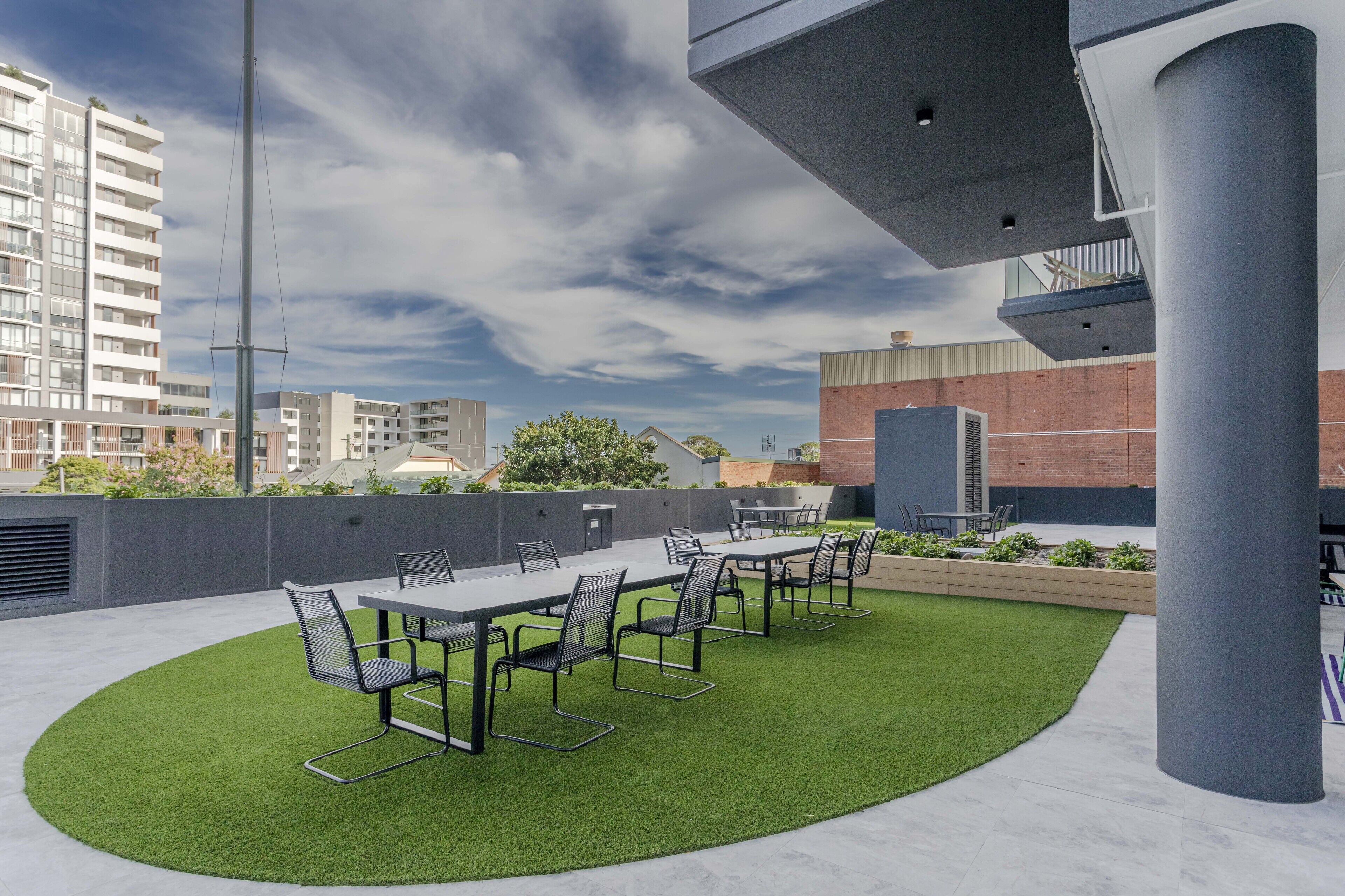 Patio esterno con un tavolo lungo e sedie, circondato da erba sintetica, sotto un edificio moderno con cielo sereno.
