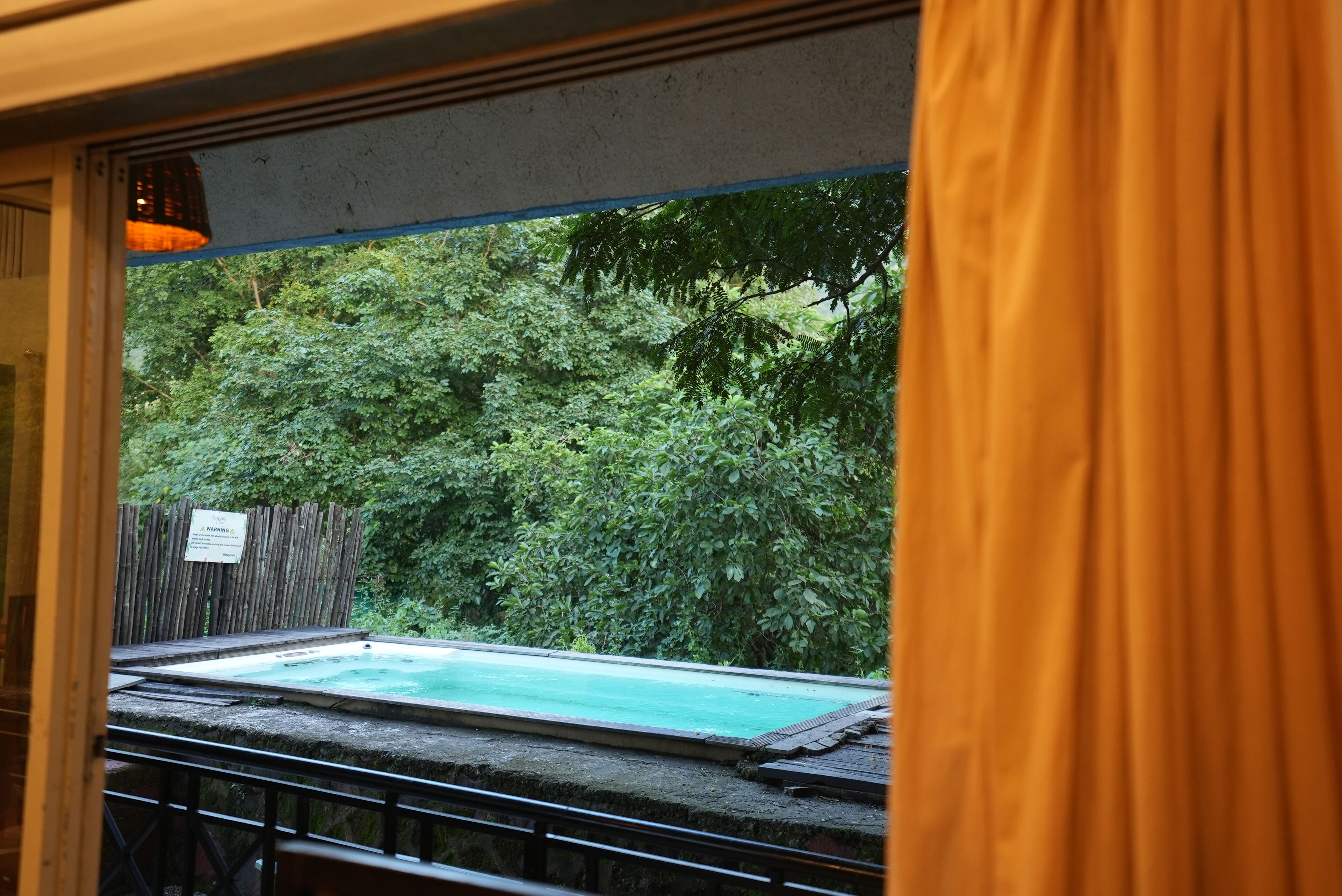 A view of a swimming pool surrounded by lush greenery, with a wooden fence and a sign in the background.