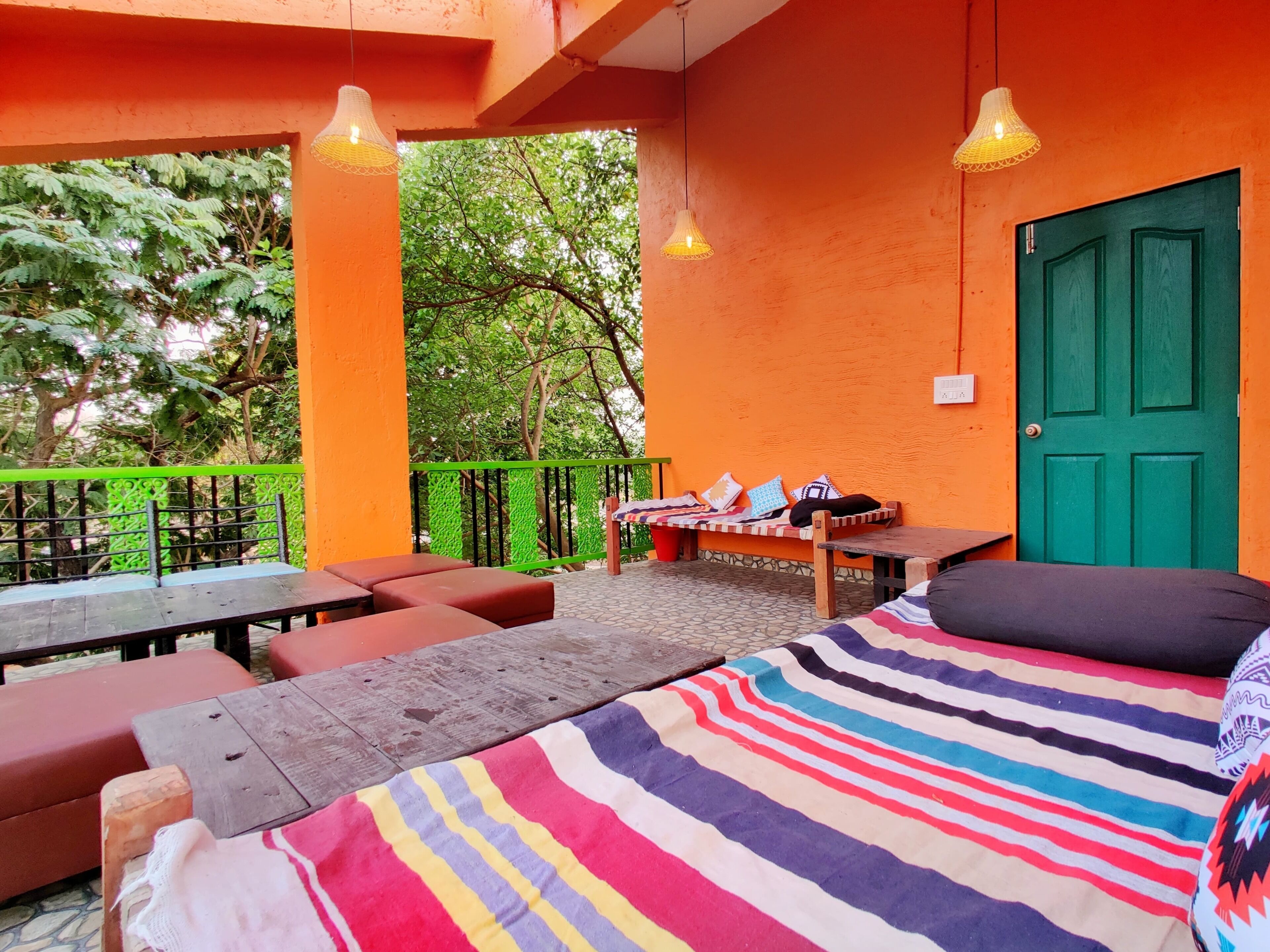 A room with a bed, a bench, and a table. There is a green door and a window overlooking trees.
