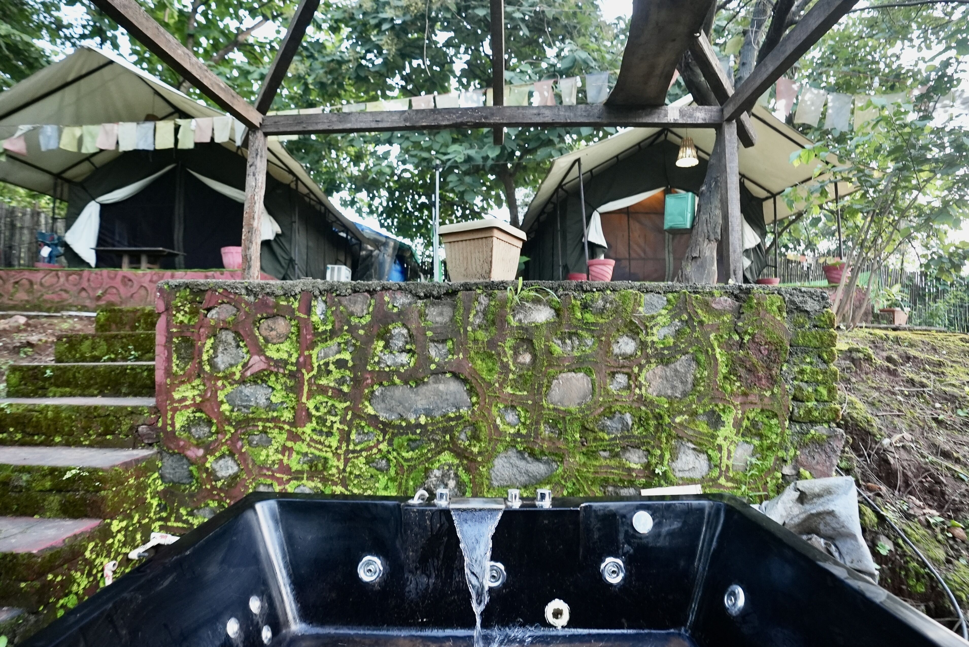 A stone wall with a black metal structure, possibly a water feature, in a natural setting with trees and tents in the background.