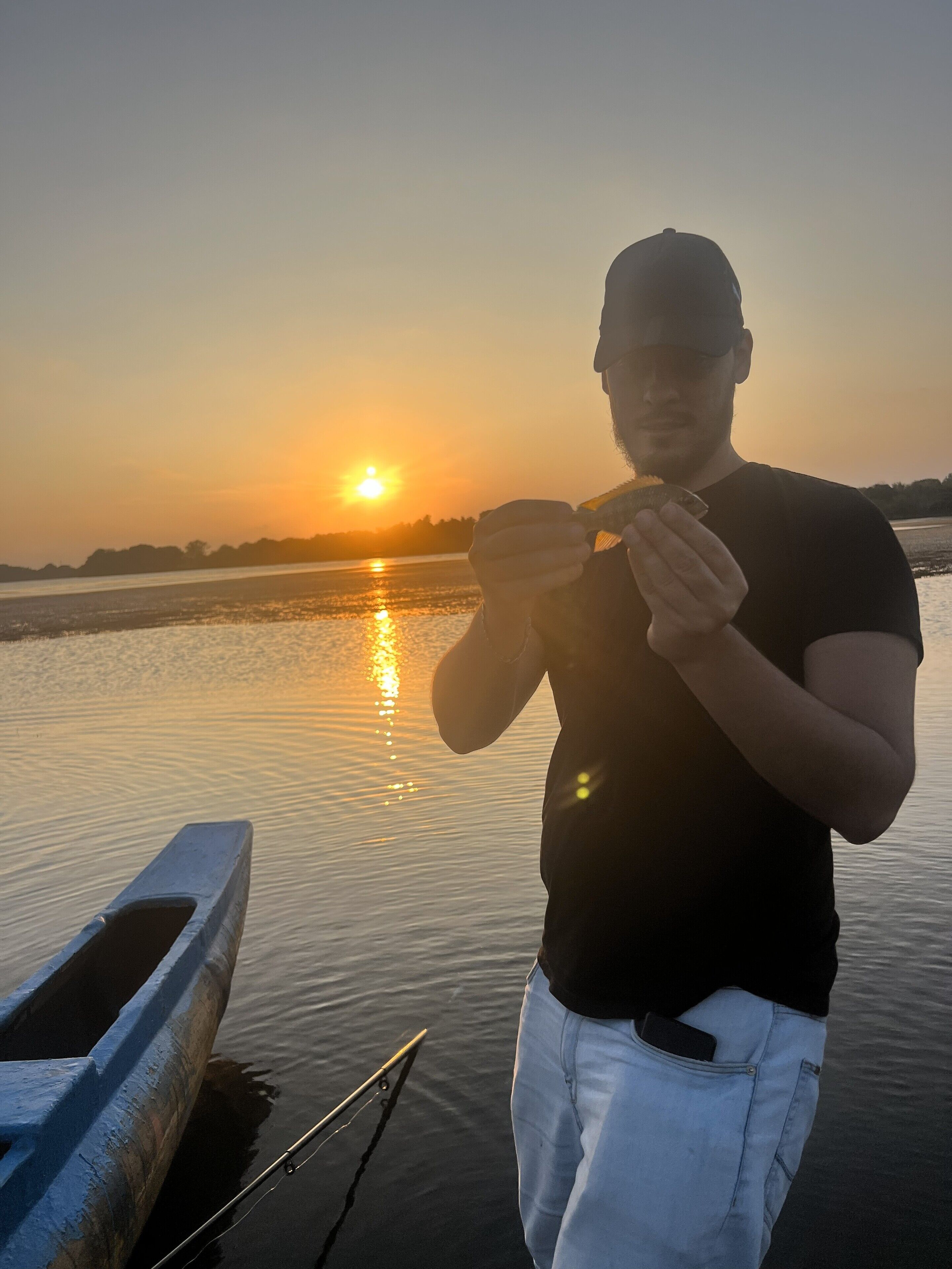A person standing by a calm body of water during sunset, holding a fish.