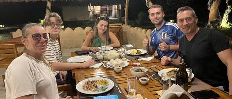 A group of people dining at an outdoor restaurant with a thatched roof, wooden tables, and a variety of dishes on the table.