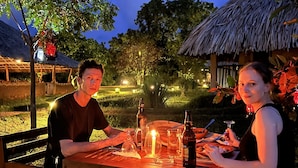 Deux personnes dînent à une table en plein air, sous un toit de chaume, entourées de verdure et sous un ciel étoilé.
