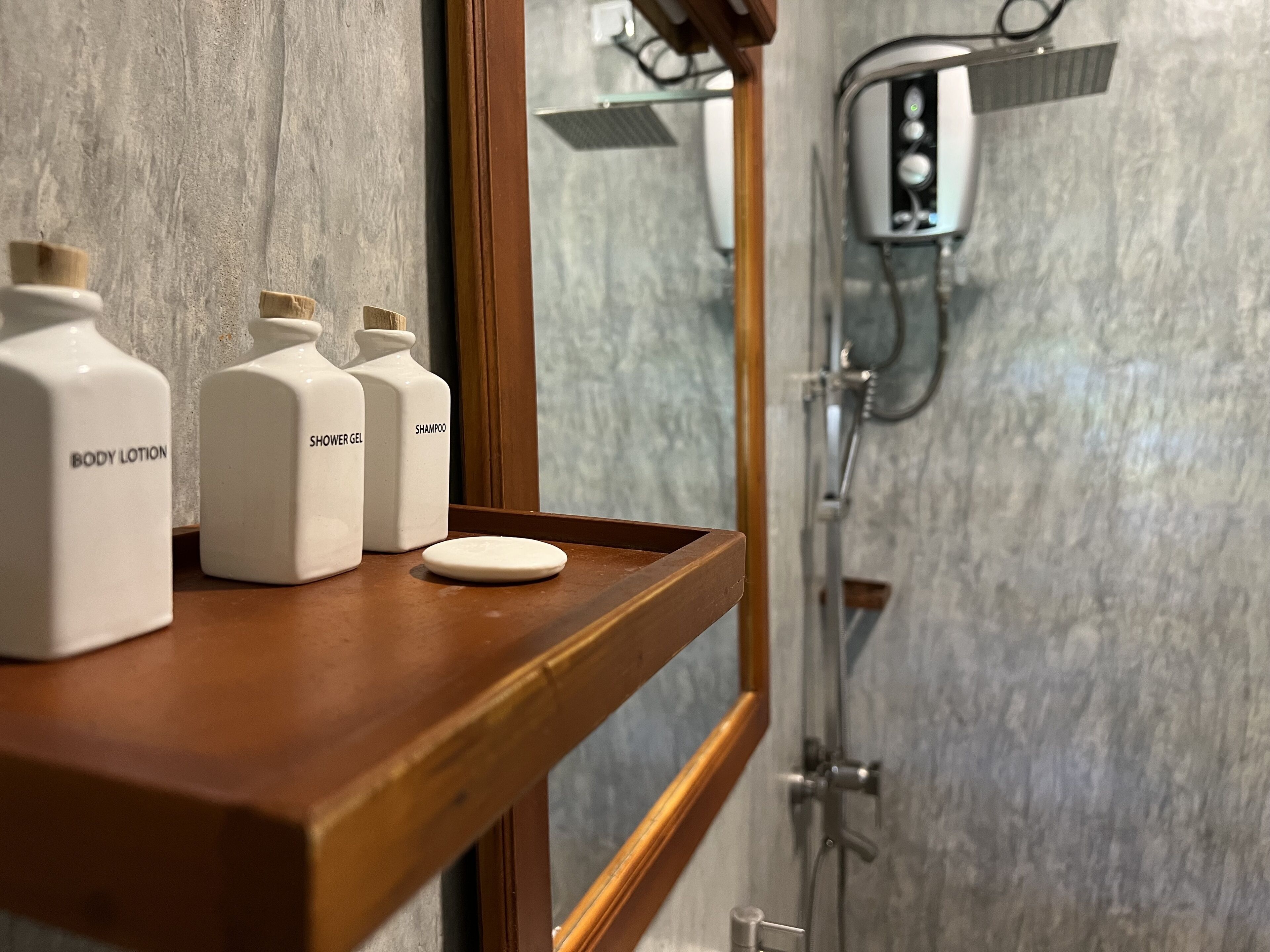 Un baño con un estante de madera que contiene tres botellas blancas etiquetadas con loción corporal, gel de ducha y champú. También se aprecia un jabonera redonda en el estante.