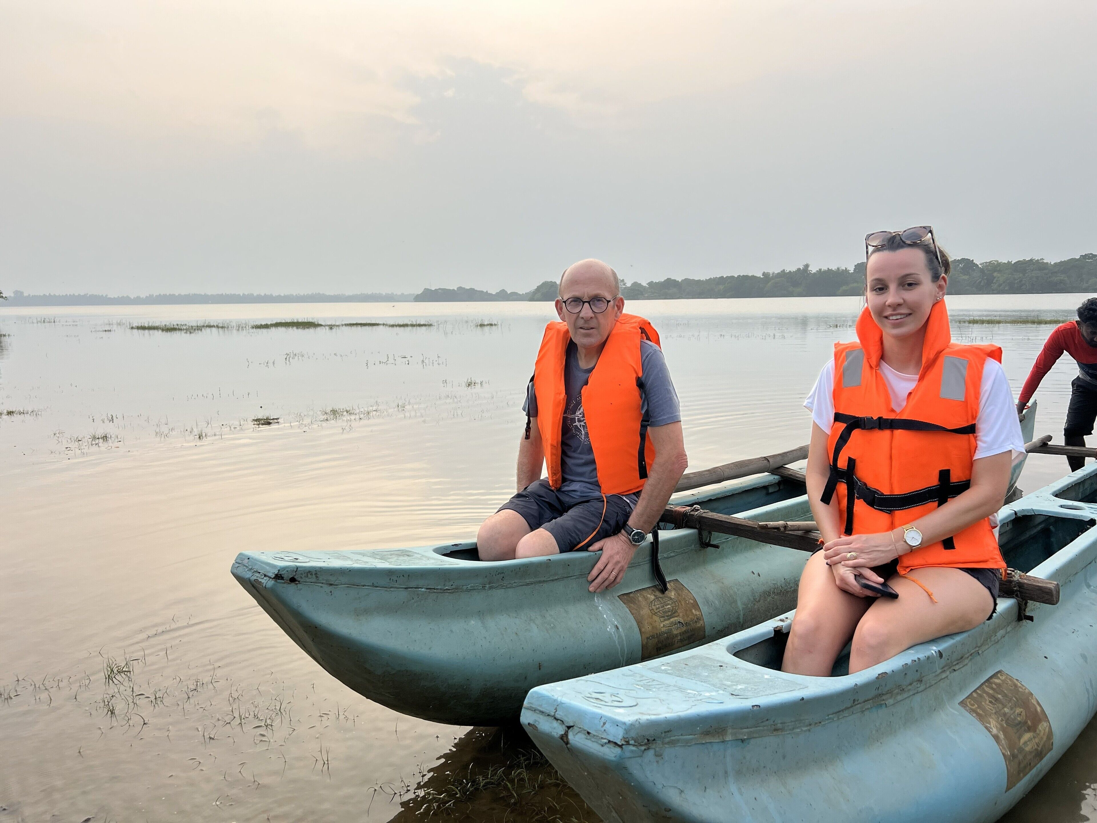 Two people in a small, narrow boat with a calm water body and distant shoreline in the background.