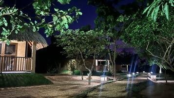 Vista nocturna de un resort con un edificio de techo de paja, un camino de tierra y árboles.