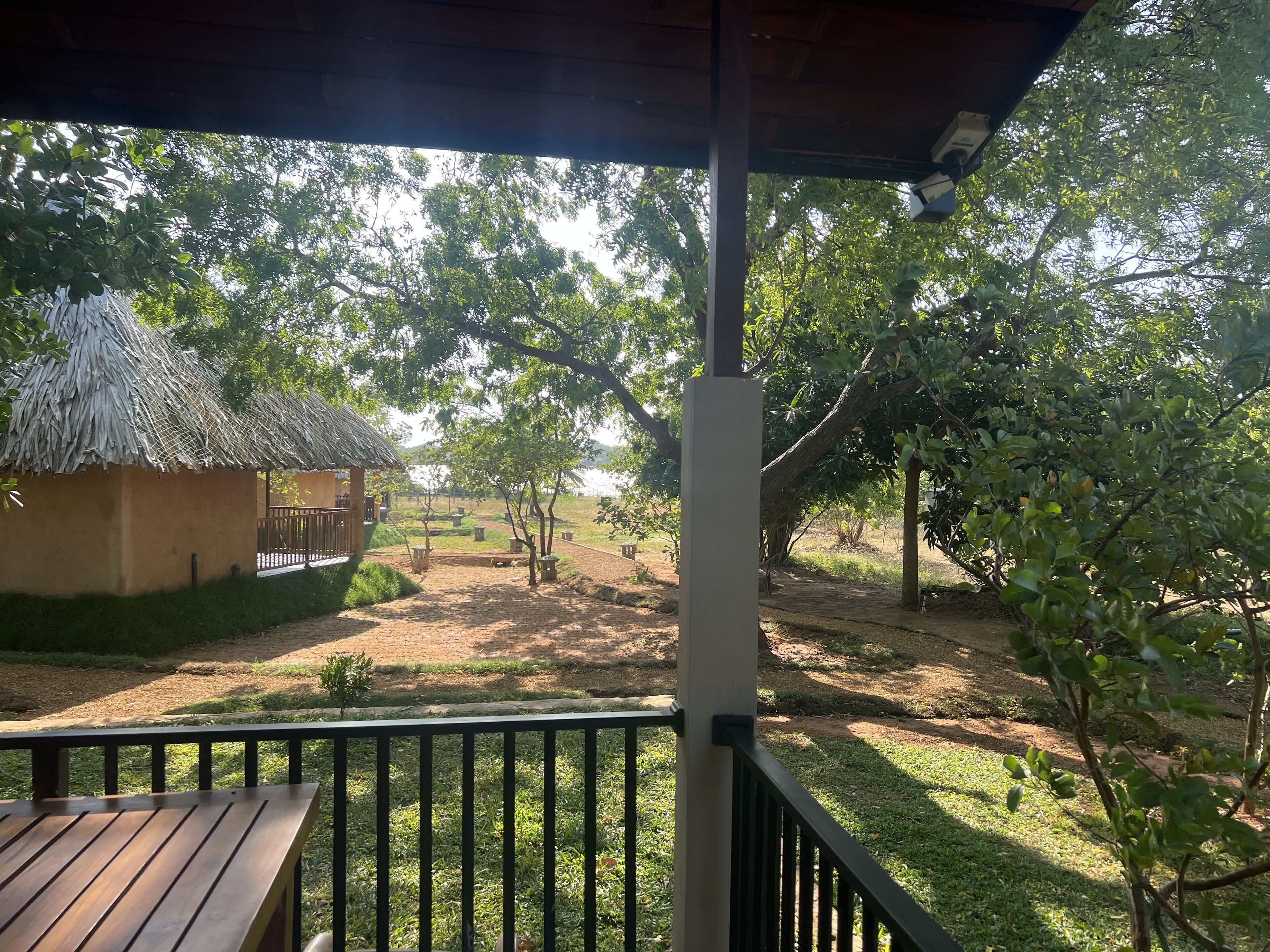 Un espacio al aire libre techado con un banco de madera, una construcción de techo de paja y vistas a un jardín con árboles y un sendero de tierra.