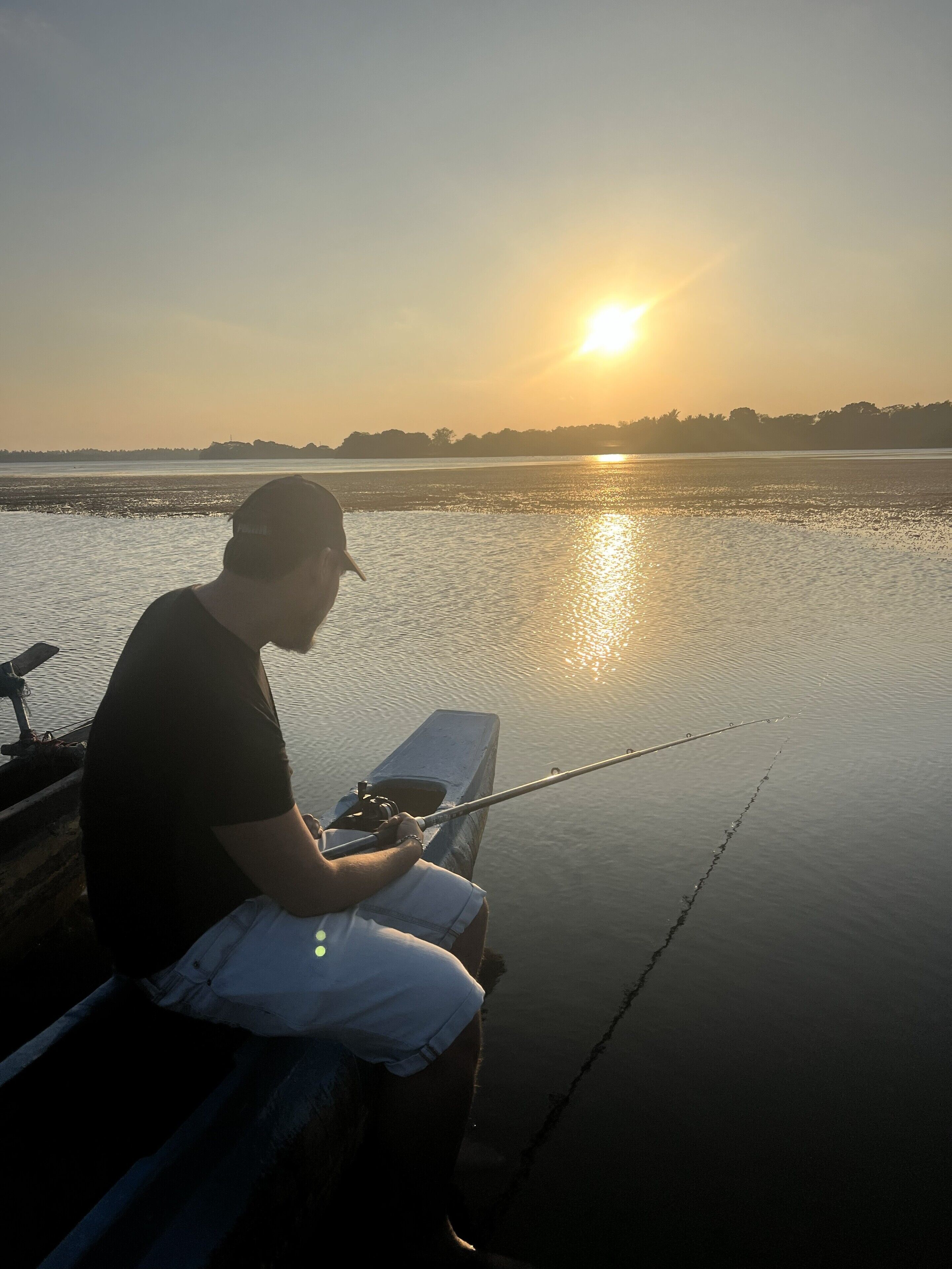 Iemand zit op een steiger met een hengel in de hand, met een rustig wateroppervlak en een ondergaande zon op de achtergrond.