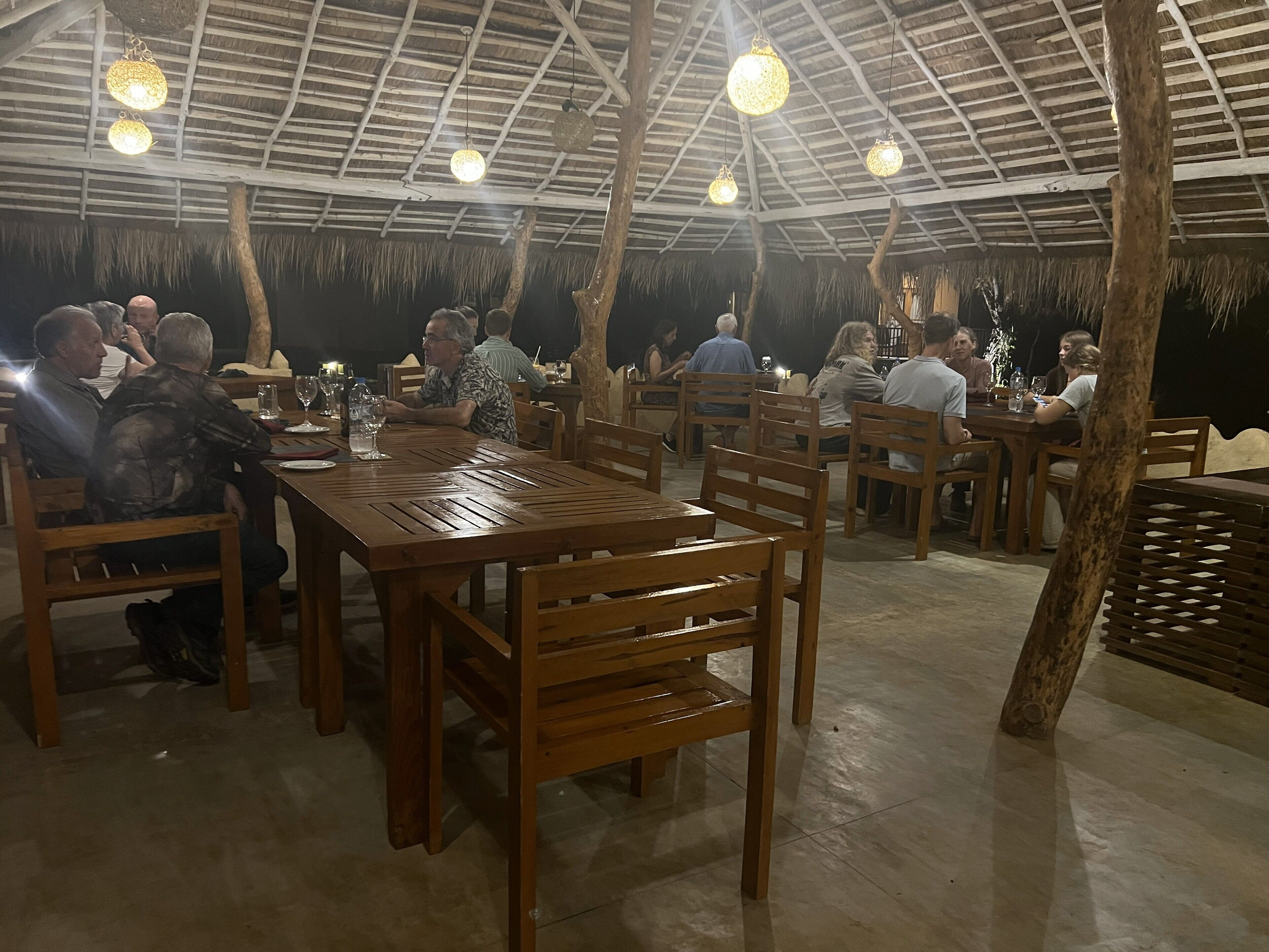 An outdoor dining area with wooden tables and chairs, a thatched roof, and hanging light fixtures.