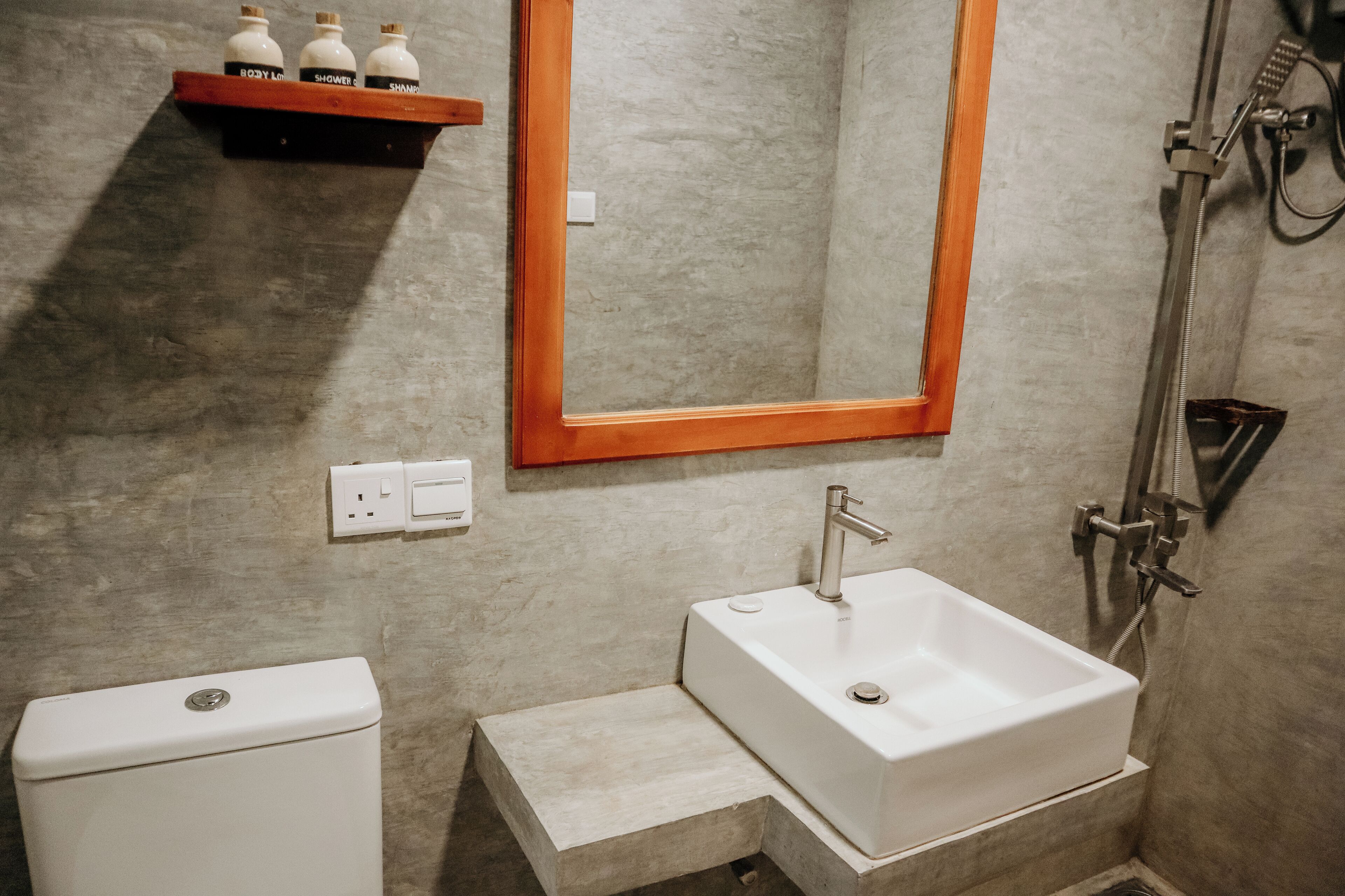 Un baño con un lavamanos blanco, un espejo con marco de madera, un cabezal de ducha y un estante con botellas.