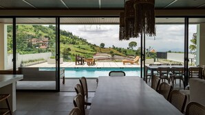 A modern dining area with a pool view, large windows, and outdoor seating. -  JadenHill CityView 8BR Villa Heated Pool+Movieroom (Cimenyan)