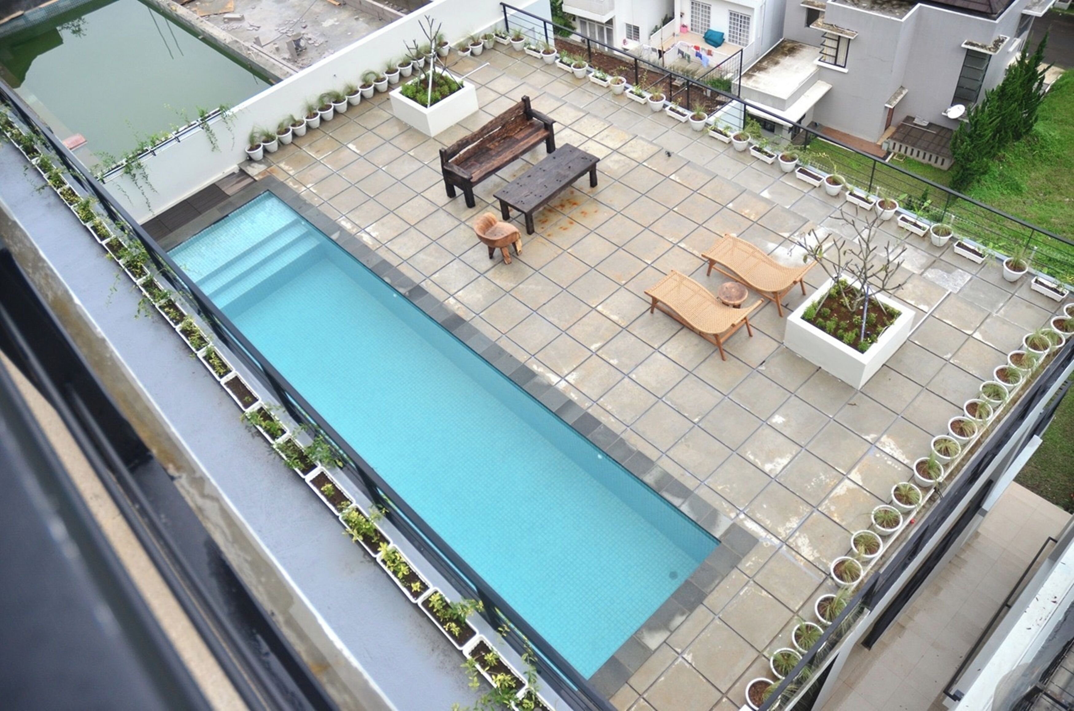 Un espace piscine aménagé sur le toit, comprenant une piscine rectangulaire, un banc en bois, une table avec des chaises et des plantes en pot disposées tout autour.