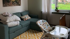 Un salon avec un canapé vert, un tapis à motifs jaunes, une poussette et un tableau représentant un paysage de village.