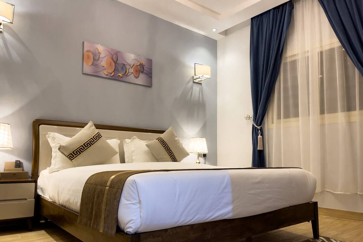 Un letto ordinato con struttura in legno, biancheria bianca e cuscini decorativi. Un comodino con una lampada, una luce a parete e un quadro sopra il letto.