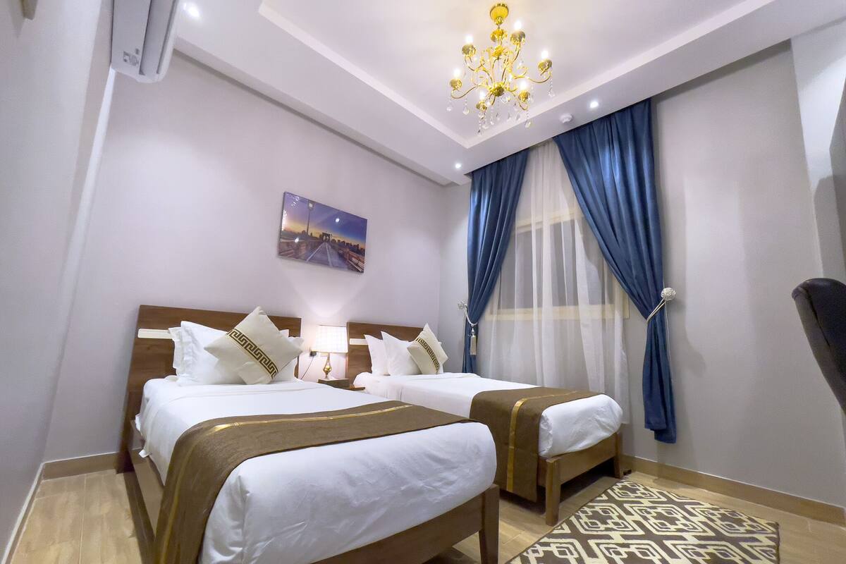 Camera d'albergo con due letti, un lampadario, un quadro e tende azzurre.