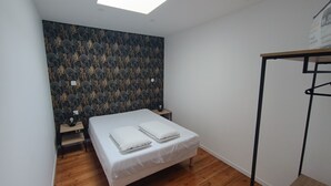 Une petite chambre à coucher moderne, avec un lit, un plancher en bois, un luminaire au plafond et un mur orné d’un motif de feuilles.