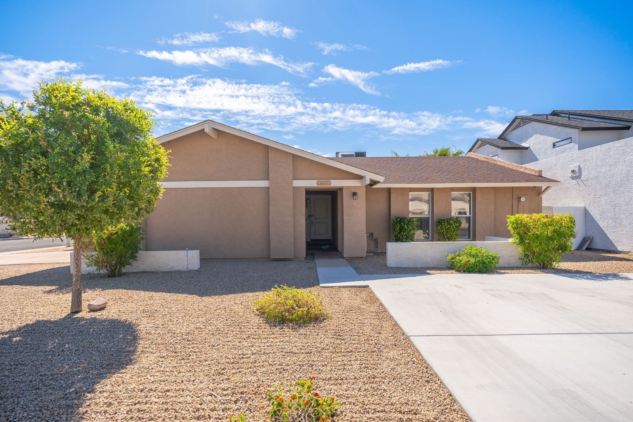 A modern single-story house with a beige exterior, a front entrance, and a small front yard with gravel and a tree.