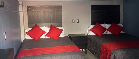 A hotel room with two beds, each with a red and white pillow, a grey quilted bedspread, and a black nightstand between them.