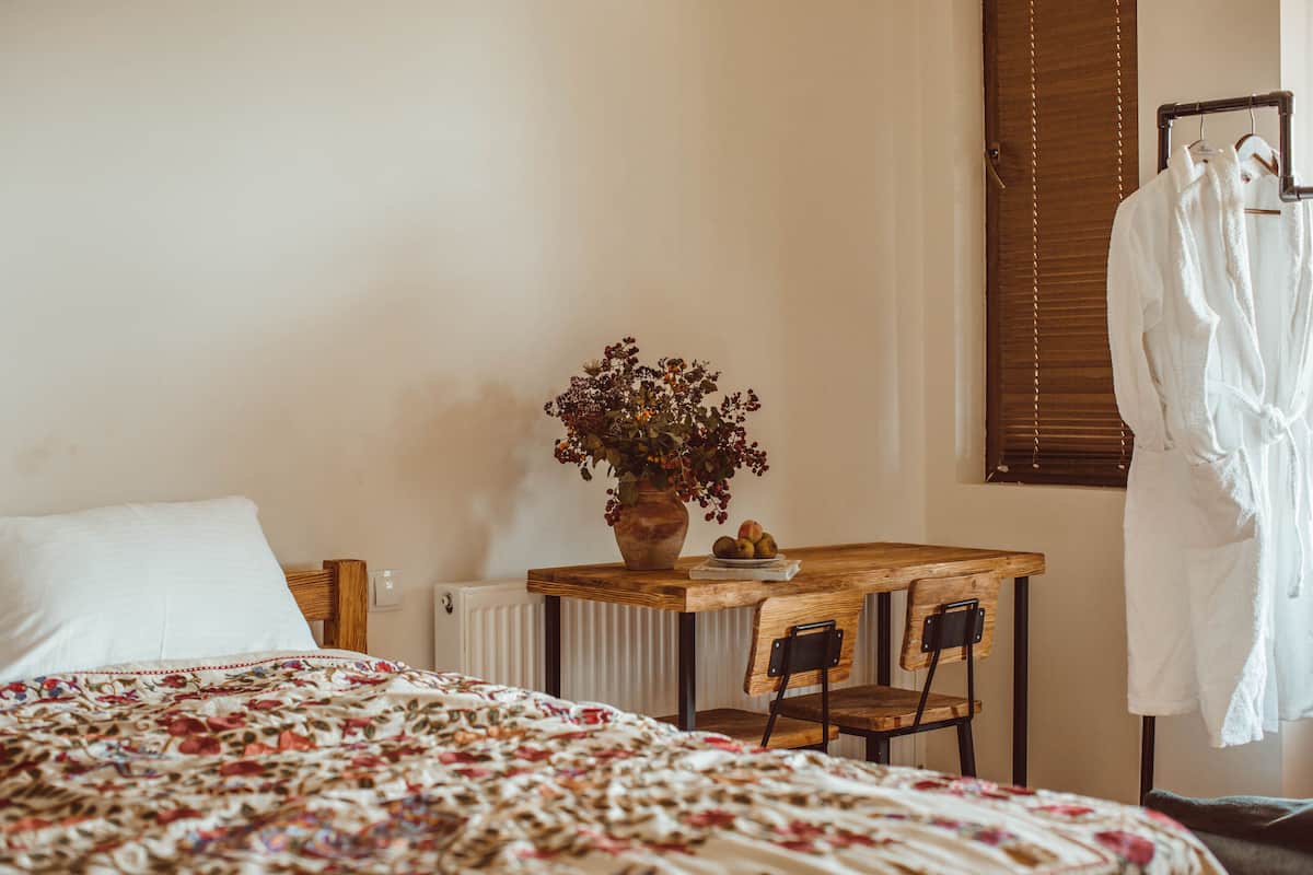 Un dormitorio con una cama, una mesa de madera con un jarrón de flores, una silla y una bata colgada en un perchero.