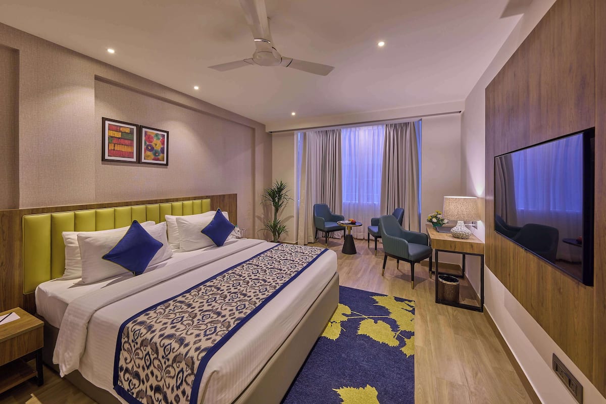 A hotel room with a large bed, a desk with a TV, a chair, a small table, and a plant.