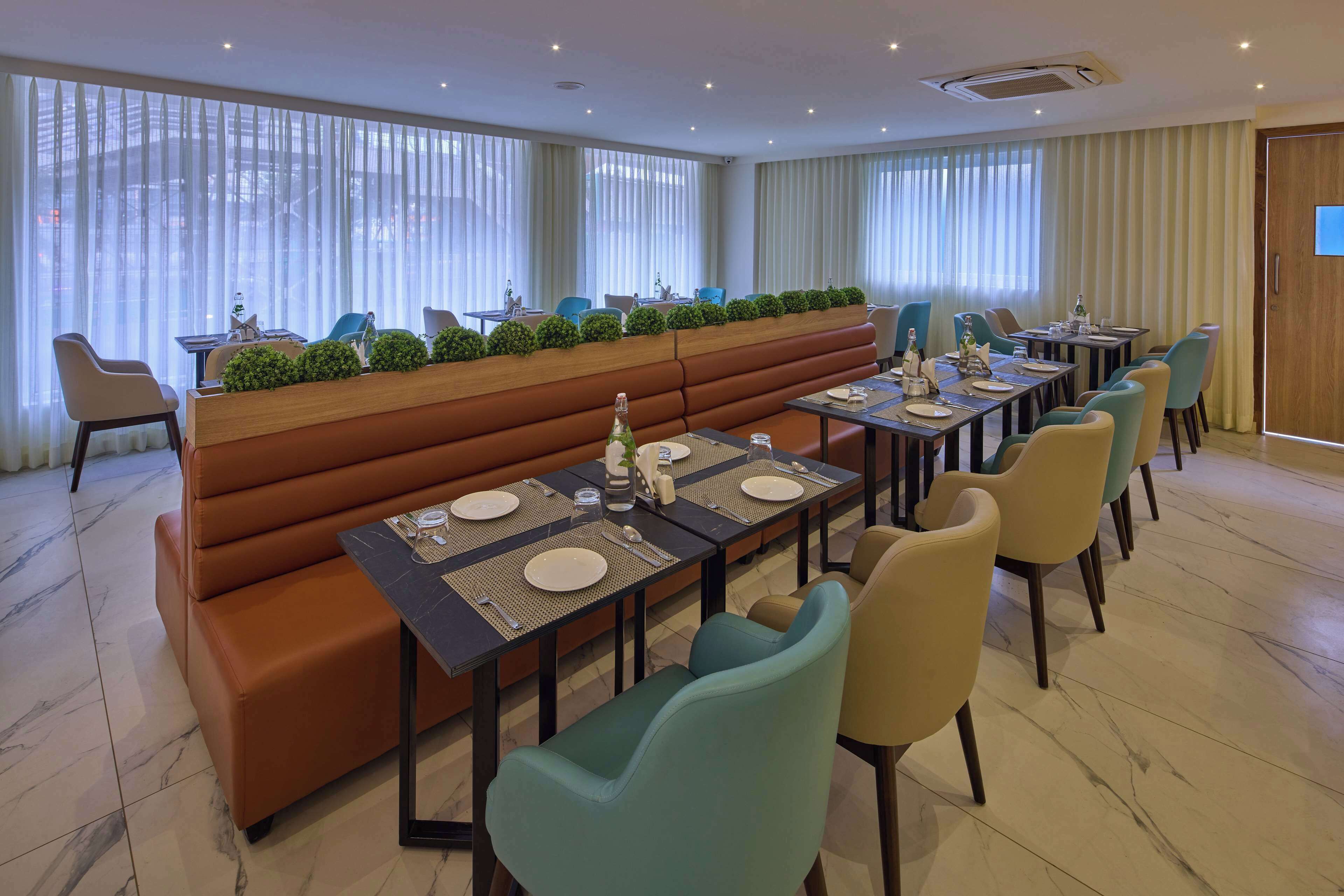 A dining area with a long table set for a meal, surrounded by cushioned seating and a row of plants.