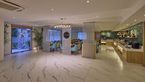 A modern hotel lobby with a marble floor, a central seating area with light blue chairs, a buffet counter with various food items, and a large potted plant. - Grand Continent Hebbal a Sarovar Portico Affiliate Hotel (Bengaluru)