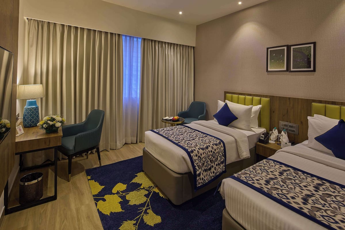 A hotel room with two beds, a desk with a lamp, a chair, and a small table with a fruit bowl.