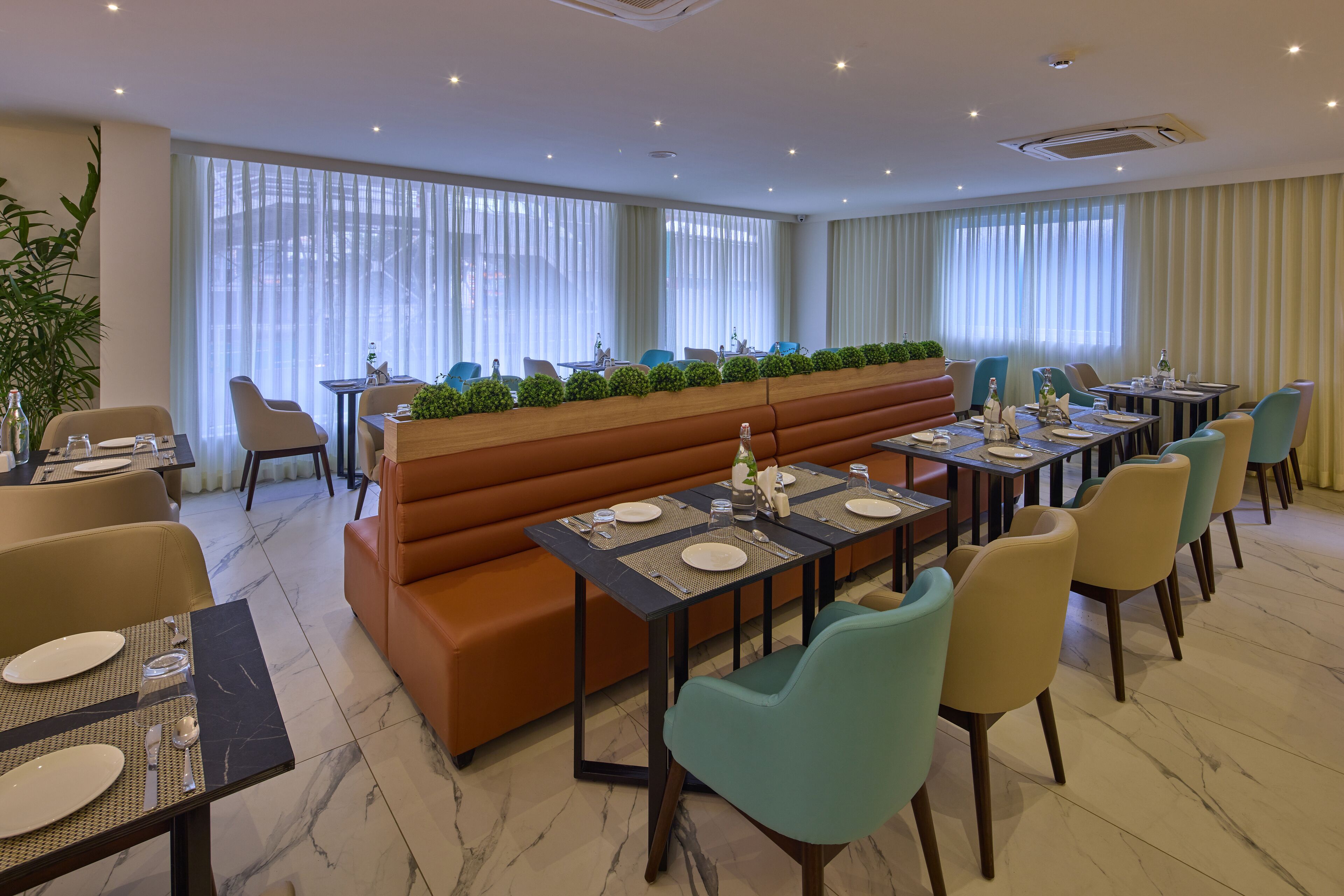 A hotel dining area with multiple tables, cushioned seating, and large windows with curtains.