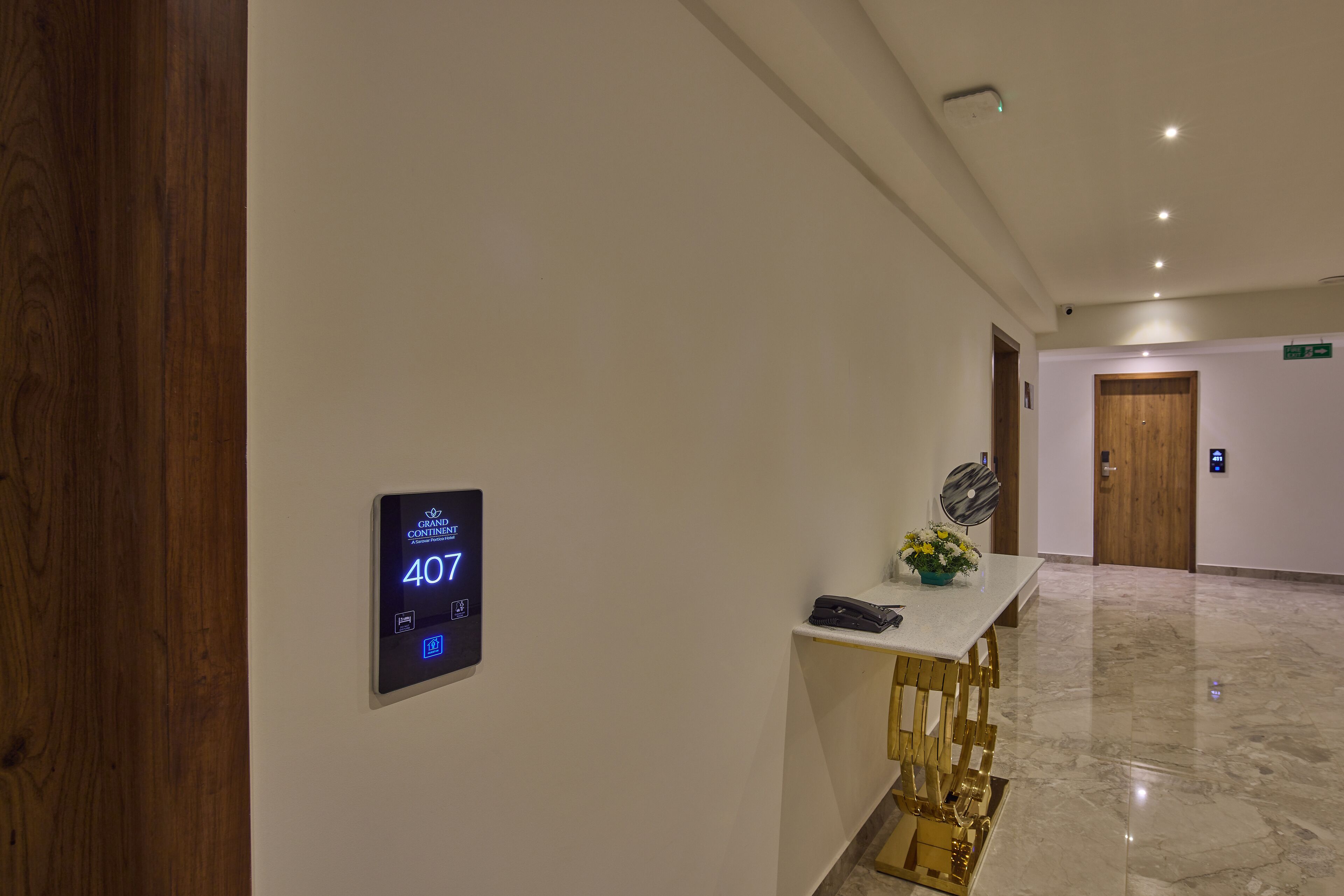 A hotel reception area with a digital display showing room number 407, a reception desk with a telephone and a vase of flowers, and a wooden door in the background.