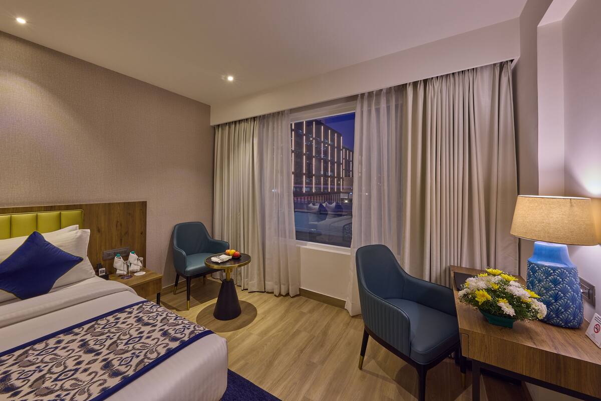 A hotel room with a bed, two chairs, a desk with a lamp, a small table with fruits, and a window with curtains.