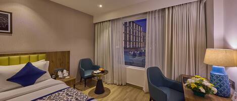 A hotel room with a bed, two chairs, a desk with a lamp, a small table with fruits, and a window with curtains.