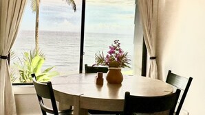 A dining area with a round table, chairs, a hanging light fixture, and a view of the ocean through large windows. - NEW:  Oceanfront, Newly Remodeled 2 bedroom condo,  WiFi, AC  (Princeville)