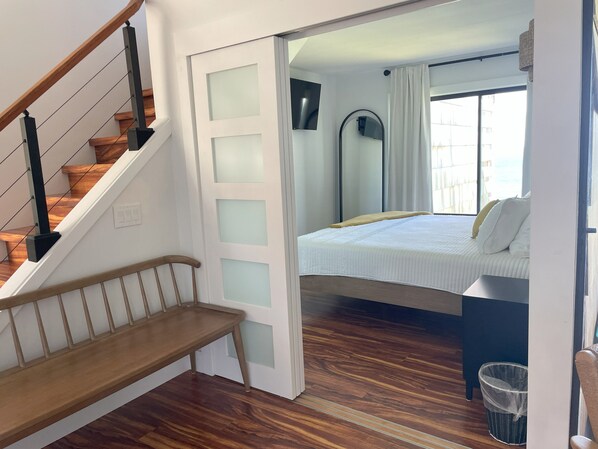 A bedroom with a wooden staircase, a bed with white bedding, a window with curtains, and a small trash bin. - NEW: Oceanfront, Newly Remodeled 2 bedroom condo, WiFi, AC (Princeville)