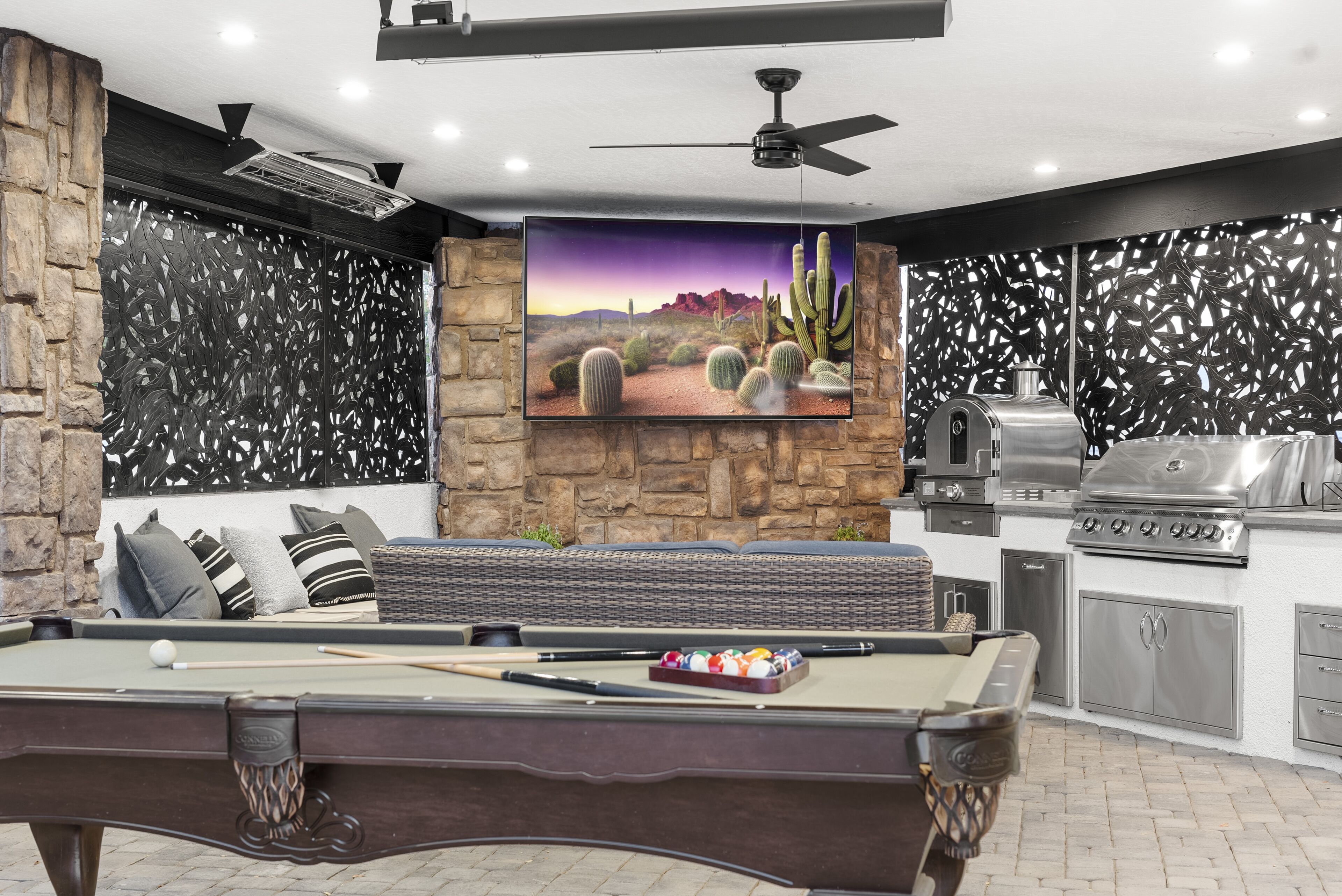 A pool table with a pool cue and balls, a sofa with cushions, a television mounted on a stone wall, and a grill area.