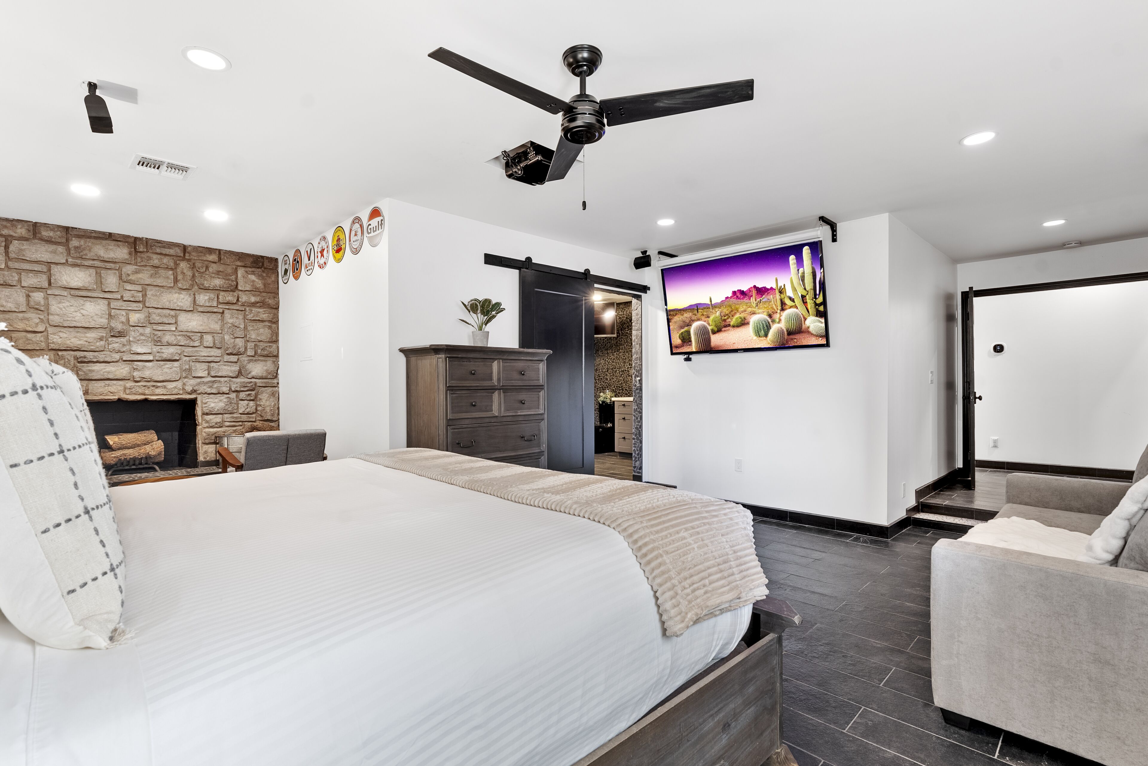 Un dormitorio moderno con una cama grande, ventilador de techo, chimenea de piedra, televisor y puerta corredera que da a un baño.