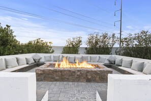 A stone fire pit surrounded by a white outdoor seating area with cushions.